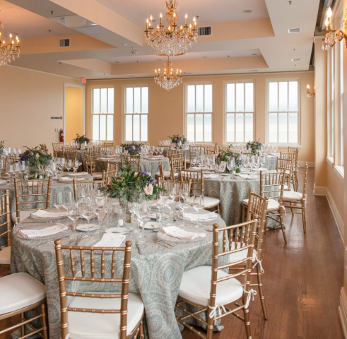 Elegant layout for a wedding rehearsal dinner in the Bullock Ballroom Elegant gold Chiavari chairs surround tables draped in blue and white linens. Tables are set with fine white china and crystal goblets. Centerpieces of hydrangeas, roses, and ferns are placed on each table.