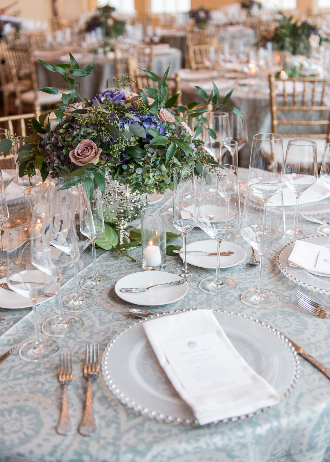 Elegant gold Chiavari chairs surround tables draped in blue and white linens. Tables are set with fine white china and crystal goblets. Centerpieces of hydrangeas, roses, and ferns are placed on each table.