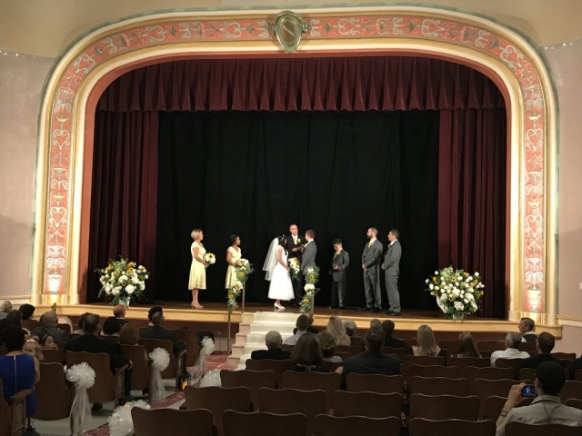 A bride and groom stand on the stage of our vintage Vaudeville theatre with several groomsmen and bridesmaids on either side. The stage is adorn with white flower and yellow sunflowers in large arrangements. Guests look on from the comfort of the theatre's comfy chairs.